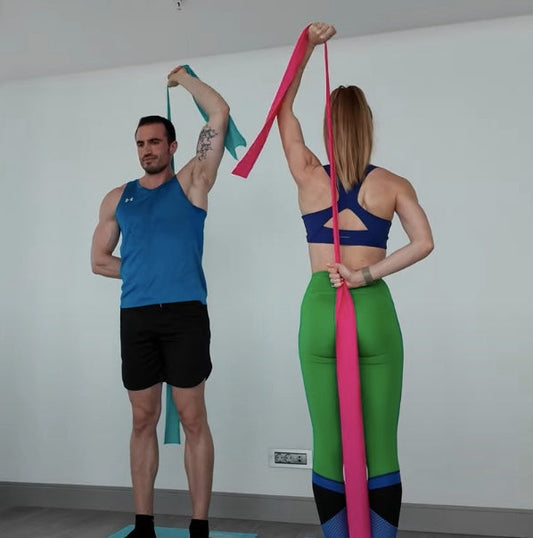 Two people demonstrating single-arm overhead triceps extension at home using resistance bands anchored behind their backs — Evde, direnç bandını sırtına sabitleyerek tek kol overhead triceps extension yapan iki kişi (kadın ve erkek).